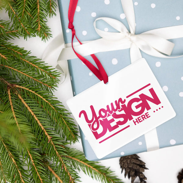 A custom acrylic ornament with a red ribbon, placed on a gift wrapped in blue and white polka dot paper, surrounded by green pine branches and pine cones.